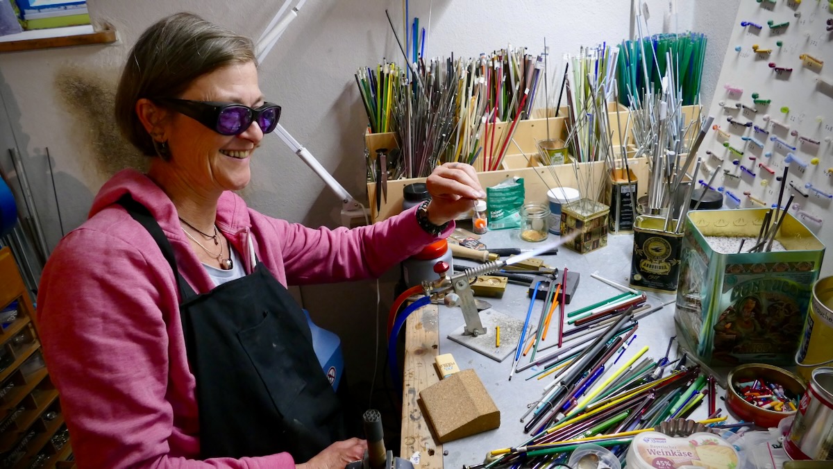 Angelika an ihrem Glasbrenner, lächelnd bei der Arbeit in ihrer Werkstatt voller Glasstäbe