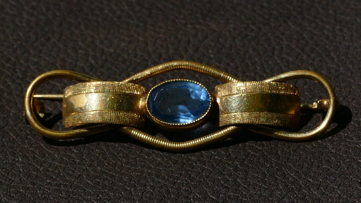 Goldene Brosche mit zentralem blauen Edelstein, fein ziseliert und gewunden, auf dunklem Untergrund. Ein Tropfen Nachtblau, gefasst in goldene Linien – wie ein Gedanke, der nicht vergeht.