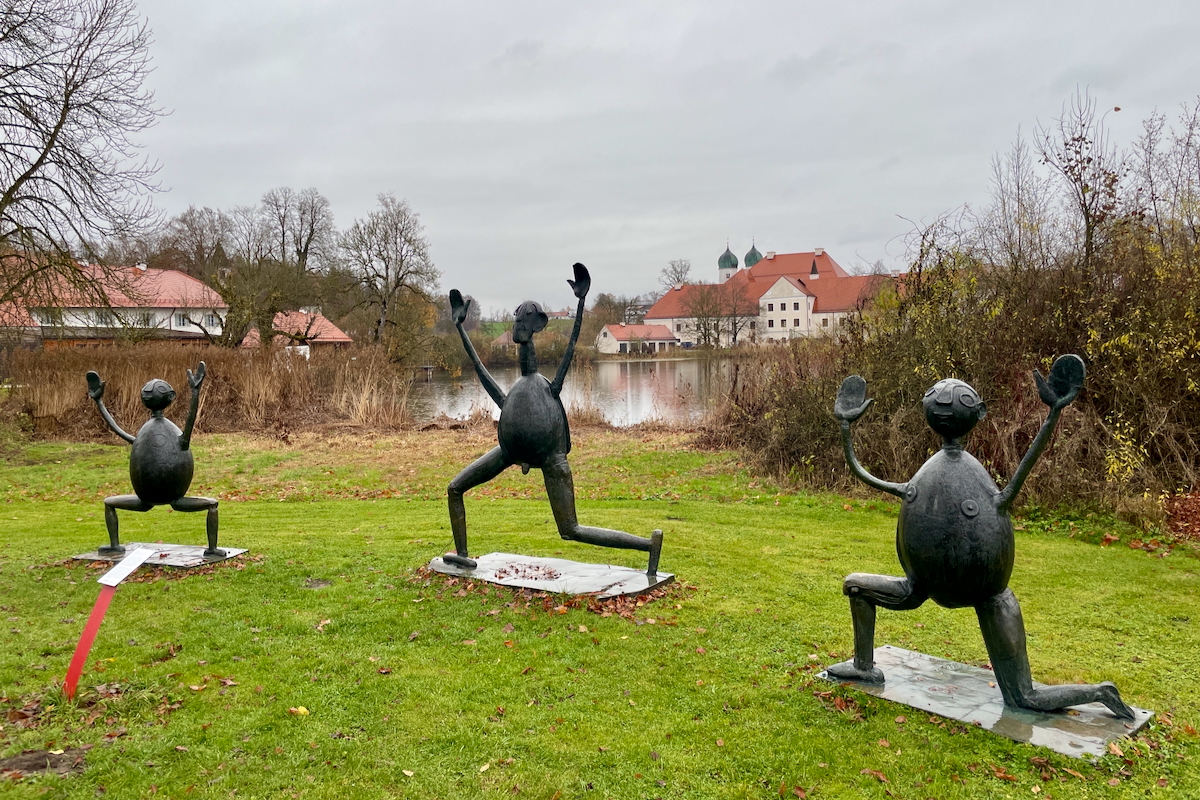 Drei humorvolle Bronzeskulpturen mit runden Körpern und erhobenen Armen stehen auf einer Wiese vor einem See und einem historischen Gebäude mit roten Dächern.
